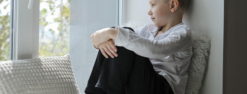 46481738 lonely sad serious school boy sitting on a windowsill in the house looking at window sunny weather